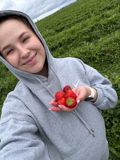 A young woman with freckles is taking a selfie in a strawberry field. She is wearing a grey hoodie and holding a handful of strawberries. The field is lush and green, with a cloudy sky overhead. The location is the city of Berkuty, Russia.