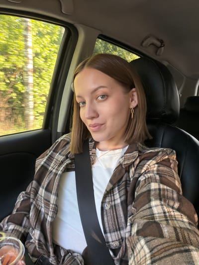 A young woman is sitting in a car. She has short brown hair, fair skin, and is wearing a seatbelt, a white shirt, and a brown and white plaid jacket. Outside the car window, trees and greenery are visible, suggesting a rural or suburban environment. Based on the specified location, the surroundings could be a drive through or near Maksimkovo, Russia. The car's interior is visible, including a black leather seat.