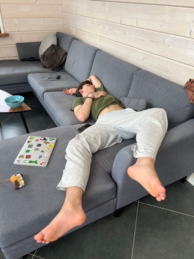 A young man is lying diagonally across a gray sectional sofa. He is wearing a green t-shirt and gray sweatpants. His arms are positioned near his head, seemingly resting or in a relaxed pose. A laptop covered in stickers is on the sofa near his feet. A small wooden toy is also next to the laptop. The floor is partially visible, showing a black tiled surface. The adjacent wall and ceiling are lined with white wooden planks. A small side table with a blue bowl on it is also visible next to the sofa. The city of Svetloe, Russia, is not identifiable in this image.
