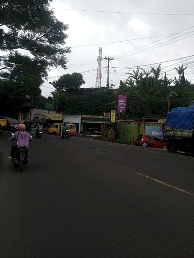 The scene depicts a street in Thanthirimale, Sri Lanka, under an overcast sky. A dark asphalt road dominates the foreground and mid-ground, showing minor surface irregularities and a faded yellow broken line on the right side. On the left, several commercial structures are visible, including a building with a yellow and pink painted facade and another with a partially obscured sign reading "GOWR KANNA." Behind these buildings, a dense expanse of green foliage, including large trees and smaller bushes, extends up an embankment.
Traffic includes multiple motorcycles and vehicles. In the immediate foreground, a person wearing a light purple hooded garment and a pink helmet rides a dark scooter away from the viewer. Further ahead on the left, two more individuals are visible on motorcycles. A black SUV is partially parked on the far left. A compact yellow and pink vehicle, possibly a Bajaj Qute, is positioned near the commercial buildings.
On the right side of the road, a red hatchback car is parked with its rear hatch open, adjacent to a green corrugated metal barrier. Behind this barrier, a billboard with "ASIA" visible is partially obscured. A large, dark-colored truck with a blue tarp covering its cargo is parked further right. Above the right-side structures and vegetation, a tall, red and white lattice-style telecommunications tower stands prominently against the cloudy sky. Utility poles with multiple power lines traverse the upper mid-ground of the image. Lush green vegetation, including banana plants, is also present on the right embankment behind the commercial elements.