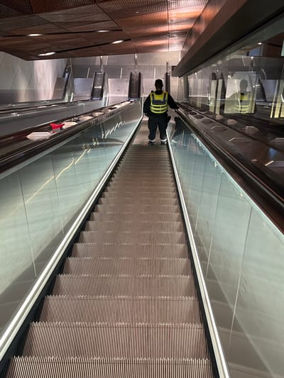 The image depicts the interior of a modern underground metro station in Espoo, Finland, specifically a Länsimetro station. The perspective is downward, looking along a very long, active escalator.

In the mid-ground, a single individual, an adult male, stands on the central escalator, descending. He is viewed from behind, wearing dark trousers, dark shoes, and a high-visibility yellow-green vest with reflective stripes and text, including "METROPOLI", printed on the back. His left hand rests on the moving handrail.

To the left of the main escalator, two additional parallel escalators are visible in the background, appearing stationary. On the handrail or side panel of the immediately adjacent stationary escalator, several white rectangular containers, resembling takeout food boxes, are present, alongside a single bright red object, possibly a utensil.

The escalators feature dark grey treads, dark handrails, and illuminated transparent or translucent side panels with integrated white LED strip lighting. The station ceiling is a distinctive reddish-brown perforated metal design with integrated lighting. Smooth, light-colored wall panels are visible behind the background escalators. The overall environment is clean, modern, and well-lit by artificial sources.