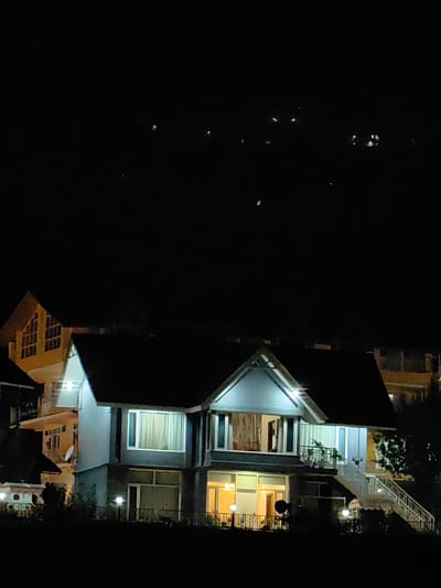 A night-time photograph captures a multi-story building with illuminated windows, suggesting interior lighting in Kulang, India. The building exhibits a contemporary architectural style, featuring prominent balconies and a set of outdoor stairs. The surrounding environment is dark, with a few scattered lights visible in the distance, possibly indicating other structures or streetlights. The sky above is black with faint, small white dots, which could be stars.