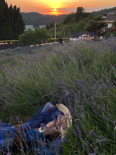 A woman lies on her back in a field of lavender, her head resting on a straw hat, with her hands near her face. She is wearing a sheer blue dress and a brown belt. Two daisies are placed on her eyelids. The lavender field extends into the background, where trees and a hillside are visible under a sunset sky. The location is identified as Verkhneye Uchdere, Russia.