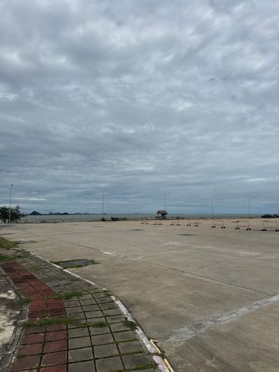 The image depicts an overcast day in Don Sak, Thailand, with a large concrete parking lot in the foreground. The lot is lined with red metal barriers with wheels, set up in rows. There are several streetlights and some parked cars visible in the distance. Beyond the parking lot, a calm body of water stretches to the horizon, with several islands or peninsulas dotting the landscape. A small wooden structure with a red roof stands near the water's edge, possibly a shelter or kiosk. The foreground features a section of paved walkway with red and grey bricks, interspersed with patches of grass. The sky is filled with heavy, grey clouds, suggesting an imminent rain or a generally gloomy atmosphere. The overall scene conveys a sense of an empty or sparsely populated public space by the sea.