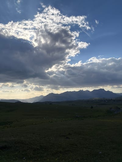 The image shows a landscape in Pišče, Montenegro. The sky is partially covered with clouds, some of which are backlit by the sun. Mountains are visible in the midground, fading into the distance with a light haze. A grassy field occupies the foreground.