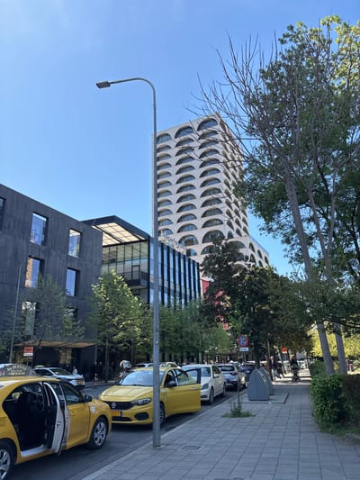 The image depicts a street in Tirana, Albania, with distinctive architectural elements. A modern building with rounded, arch-like windows dominates the left side of the frame, adjacent to a lower, darker-colored building with rectangular windows. A lamppost stands tall, partially obscuring the view of the buildings. On the right side of the street, a row of parked yellow taxi cabs is visible. The cabs are parked along a paved sidewalk. Trees are present along the street, providing some shade. In the background, there are hints of other buildings and people walking. The sky is clear and blue, suggesting a sunny day.