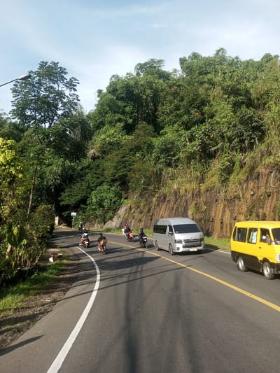 The image depicts a road scene in Phou Khoun, Laos. The road curves slightly to the left and has a yellow dividing line. A silver van and a yellow car are driving on the road. Several motorcycles are also present. Lush green vegetation covers the hillside adjacent to the road. The sky is partially cloudy. A street lamp is visible on the left side of the road.