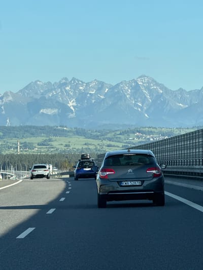 The image presents a view from a highway in what appears to be a mountainous region. Cars are traveling on the road. In the background, there is a panorama of mountains, with slopes covered in green vegetation and rocky peaks. The sky is clear and blue. The license plate on the car closest to the camera reads "KWA 52878." There is an overpass to the left and a white railing to the left side as well. Given the "PL" on the license plate, and the image context, it is likely near Lasek, Poland.