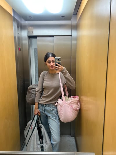 The scene depicts a female individual taking a mirror selfie inside an elevator, located in Aldoar, Foz do Douro e Nevogilde, Portugal. The elevator features brushed metallic stainless steel doors and a left wall panel, with a contrasting wooden or wood-paneled right wall. The ceiling contains a recessed rectangular light fixture with two visible light sources. The floor is a light-colored, possibly marble, surface.
The subject, reflected in the mirror, is a female with dark hair styled in an updo. She is wearing a light brown, ribbed, long-sleeved top, light blue denim jeans, a thin gold-colored necklace, and a gold-colored bracelet on her left wrist. She holds a smartphone with a clear case in her right hand, pointed towards the mirror to capture the image.
She is carrying three distinct bags: a pink, quilted, padded shoulder bag hanging from her left shoulder; a large, greyish-brown, soft duffel bag slung over her left shoulder and positioned behind her; and a light grey fabric bag with dark green trim held in her left hand. The elevator doors are closed, and a red indicator light is visible on the left side of the metallic door frame.