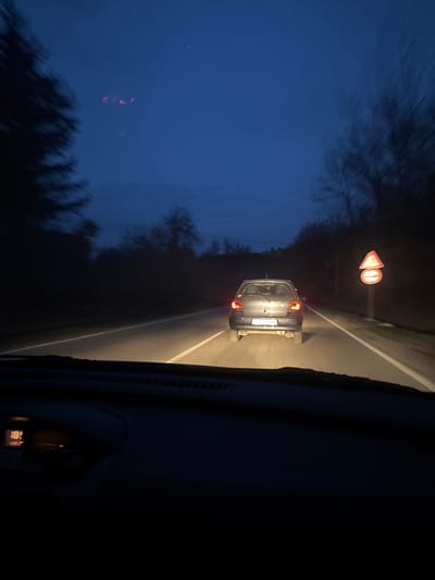 The scene depicts a late evening or night drive from inside a vehicle on a road in Rahovtsi, Bulgaria. A dark grey compact car, possibly a Toyota Yaris, is traveling ahead in the same lane, its red taillights illuminated. The asphalt road is visible, marked by a solid white line on the right shoulder, illuminated by the headlights of the observing vehicle. On the right side of the road, approximately 20 meters behind the lead car, two circular road signs are mounted vertically on a single pole: an upper red-bordered triangle with an unclear symbol and a lower red circle with a white background and a horizontal black bar (No Entry). Both signs are reflective. Dense, dark foliage and trees line both sides of the road, appearing blurred due to motion. The sky above is a dark, twilight blue. The dashboard of the observing vehicle is visible at the bottom of the frame, with an amber digital display showing "18" on the left side, likely indicating temperature. Red reflections are faintly visible on the windshield in the upper left.
