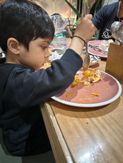 An indoor scene in a restaurant in Kalyan-Dombivli, India, depicts a young child seated at a light-colored wooden table. The child, with dark hair, is seen in profile from the chest up, facing right, wearing a black long-sleeved garment. A black thread bracelet is visible on his right wrist. He holds a silver-colored spoon in his right hand, positioned over a round pink plate with a white rim, which contains remnants of a multi-colored food item (white, green, orange/red) and reddish spice residue. To the left of the plate, a clear glass of water with condensation and a crumpled white paper napkin are on the table. In the background, a person with a light green shirt is visible holding a clear plastic water bottle to their mouth. To the right, another person's arm and upper torso are visible, wearing a dark t-shirt with partially legible white text, and orange-painted fingernails. A wooden caddy holding additional silver utensils is situated further right on the table. Other similar pink plates, mostly cleared, are also visible. The background includes blurred green foliage, suggesting decorative plants, and soft ambient lighting.