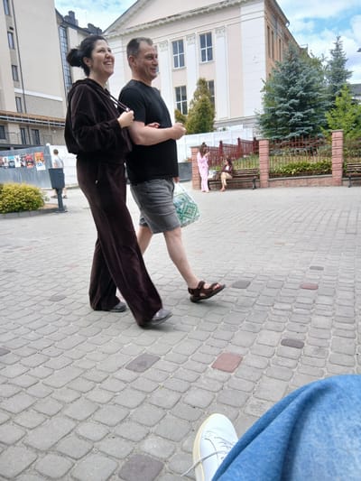 A man and a woman are walking arm in arm on a cobblestone sidewalk in Truskavets, Ukraine. The woman is wearing a brown velour tracksuit and the man is wearing a black t-shirt and grey shorts. They are both smiling and appear to be enjoying themselves. In the background, there is a building with a light-colored facade and several trees. Two women are sitting on a bench in the distance. The sidewalk is made of interlocking grey pavers with occasional red pavers.