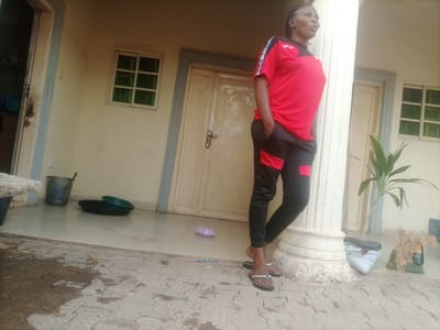 In Wuro Dole, Nigeria, a woman stands on a patterned concrete patio in front of a cream-colored building. She is wearing a red and black athletic outfit, with black pants featuring red accents. Her hair is braided and pulled back. She is looking upwards and to the left with her mouth open. To her left, there is a large white column. In the background, a cream-colored door with decorative carvings and a silver handle is visible, flanked by two windows with green panes. Various objects are present on the patio, including a grey plastic basket, two dark-colored basins (one with a green basin on top), a small purple rectangular object, and a potted plant in a dark bag. Further to the right, there is another bag with dried plant material. The patio floor shows patches of water.