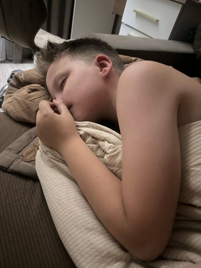 A young boy is sleeping in a bed. He is lying on his right side, facing away from the camera. His head is resting on a brown pillow, and he is covered by a beige blanket. His right arm is bent at the elbow, and his hand is near his face. The room appears to be a bedroom, with a dresser and a closet visible in the background. The lighting is dim, suggesting it is nighttime or the room is dark. There is no information to determine the specific location within Peremyshliany, Ukraine.