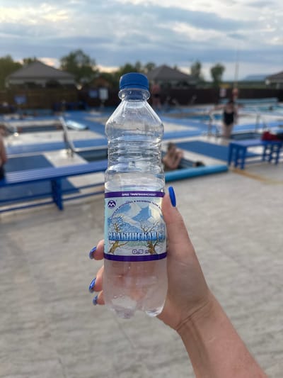 A person's hand with bright blue fingernails holds a clear plastic bottle of mineral water in front of a blurred background of an outdoor swimming pool area. The bottle has a label with text in Russian, featuring a winter landscape with mountains and a tree. The water level is about halfway down the bottle, and condensation is visible on the exterior. The background shows swimming pools with blue and white tiling, some lounge chairs, and a few people in the water or near the pools. Structures resembling small buildings or changing rooms are visible in the distance, along with trees and a cloudy sky. The lighting suggests it is late afternoon or early evening. The location is identified as the city of Yuzhnyye Koryaki, Russia, likely at a recreational facility.