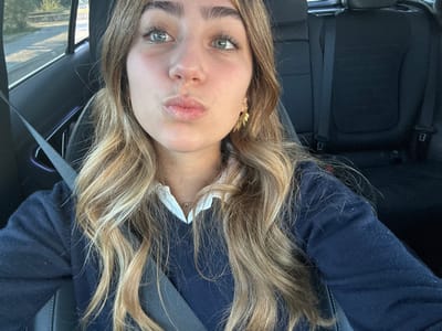 A young adult female is seated in the front passenger seat of a vehicle during daylight hours, captured in a selfie-style shot. She has fair skin, light-colored eyes, and long, wavy blonde and light brown hair. Her lips are pursed into a "kissing" gesture. She wears a dark blue V-neck sweater over a white collared shirt and a thin gold-colored necklace with a small pendant. A gold-colored earring with a leaf-like design is visible on her right ear. A dark grey seatbelt is fastened diagonally across her chest. The vehicle's interior features dark-colored seats, visible in the background rear, and dark dashboard elements. Through the window on the left, a blurred outdoor scene shows a road surface and green foliage under natural lighting. The specific location within Aldoar, Foz do Douro e Nevogilde, Portugal, cannot be determined from the visual evidence in the image.