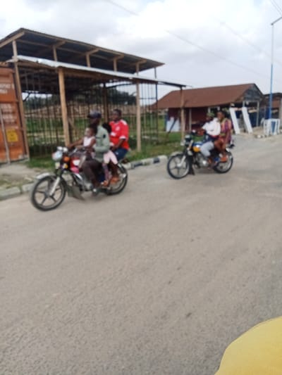 The image shows two motorcycles on a paved road in Sapele, Nigeria, at the coordinates provided. The first motorcycle, closer to the camera, has three people on it: a man driving, a woman sitting behind him, and a young girl sitting behind the woman. The second motorcycle, further from the camera, also has three people on it: a man driving, a woman sitting behind him, and a young child sitting behind the woman. Both motorcycles are traveling in the same direction, away from the camera. There are buildings on either side of the road. The building closest to the camera is a large wooden structure with a metal roof. There are power lines running overhead. The sky is partly cloudy.