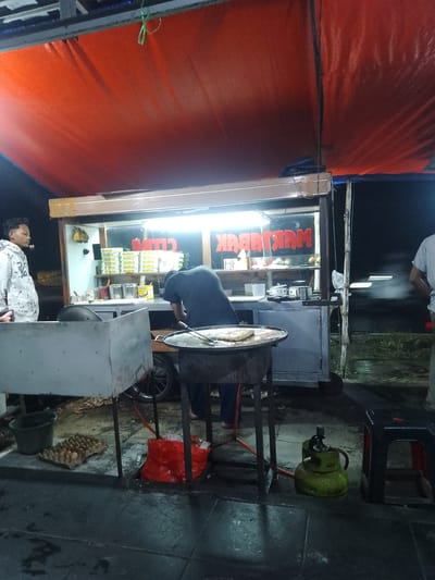 An outdoor food stall is shown at night in Jonggol, Indonesia, with a red awning overhead. A person in a white hooded sweatshirt stands on the left, facing a food cart. Inside the cart, a person in a dark shirt is preparing food on a large, flat griddle. The cart has a glass display case with yellow boxes stacked inside and a sign that reads "MARTABAK" in red lettering. In front of the cart, there is a metal structure with a cooking area, a green gas tank, and a red bag of ice. Eggs are visible in a tray on the ground to the left. The ground is wet, suggesting it has recently rained.