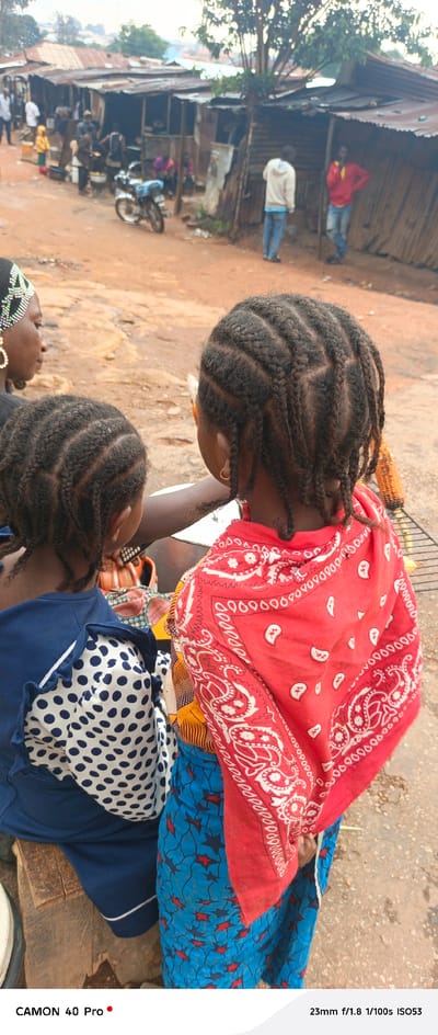 This vertical, full-frame photograph captures a scene in Barikin Gidar Kanat, Nigeria, likely in the late afternoon given the soft lighting and shadows. The foreground is dominated by the backs of three individuals. The two in the center are young girls with intricately braided hair. The girl on the right wears a vibrant red bandana with a white paisley pattern tied around her shoulders, and a blue and red patterned skirt. The girl to her left wears a blue garment adorned with white polka dots. To the left, a woman, partially visible, wears a black top and a decorative headband with beads and sequins.
Behind the individuals in the foreground, the scene opens up to a bustling street market. Makeshift stalls with corrugated metal roofs line a dirt path. Various objects are visible, including cooking pots, what appears to be food for sale, and a motorcycle. Several other people are scattered throughout the market, some in casual attire. The background reveals more of the settlement, with buildings constructed from corrugated metal and some greenery. The overall atmosphere suggests a typical day in a local Nigerian market. The visible text at the bottom of the image reads "CAMON 40 Pro" and "23mm f/1.8 1/100s ISO53", likely camera metadata.