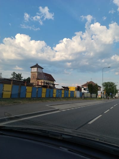 The image shows a view from a vehicle looking towards a street and buildings in Horažďovice, Czechia. In the foreground, the dashboard and windshield of a vehicle are visible. The street in front is paved with asphalt and has white lane markings. To the left of the street, a blue and yellow fence runs along a grassy area. Behind the fence, there are construction materials and a yellow structure, possibly a container or portable building, with a red crane on top of a white truck. Further back, a red-brick building with a tower stands prominently. This building features a sign with the word "Elika" in a white oval logo, repeated twice. To the right of this building, another building with a reddish roof is visible, surrounded by trees. A street lamp is positioned on the right side of the road. The sky is blue with scattered white clouds. A pedestrian with dark clothing is walking on the sidewalk on the right side of the street.