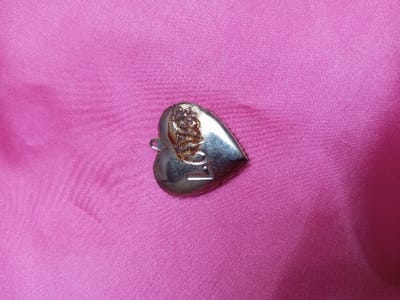 A silver-colored heart-shaped charm or pendant rests on a bright magenta/fuchsia pink fabric surface. The charm is positioned slightly off-center to the right, with its pointed bottom angled towards the lower right corner of the frame. The upper left lobe of the heart displays an irregular, dark, textured area, possibly depicting an ornate design, tarnish, or discoloration. A small, metallic loop for attachment is visible on the upper left edge of the charm. Near the bottom right point of the heart, a horizontal indentation or etched mark is discernible, potentially forming a letter "L" or "I". The metallic surface exhibits some reflectivity. The pink fabric background is largely unpatterned, showing subtle folds and variations in light reflection across its woven texture. There are no people, explicit actions, or direct interactions depicted in the scene. The image is documented in Qarabucaq qəsəbə, Azerbaijan.