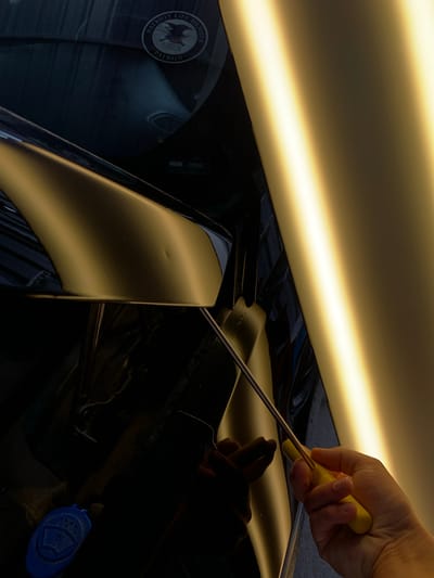 A close-up shot shows a hand holding a yellow-handled tool, likely a dent repair rod, on the side of a dark-colored vehicle with a matte gold panel. The tool appears to be inserted behind the gold panel, suggesting an attempt to repair a visible dent. In the upper left corner of the image, a circular sticker with an eagle and the text "PATRIOT LIFE MEMBER" and "PATRON" is affixed to the vehicle's surface. A bright, illuminated panel runs vertically along the right side of the frame, casting a strong yellow light and creating reflections on the car's surface. The background is dark and somewhat indistinct, showing the interior of a garage or workshop. The presence of the dent repair tool and the lighting suggest this image was taken in a professional auto body shop.