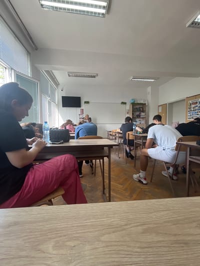 The image depicts a bright classroom scene in Nova Zagora, Bulgaria, captured from the back-right, looking towards the front-left of the room.
**Subjects:** Several students are seated at individual wooden desks with matching chairs. In the immediate foreground, the lower body of a person wearing pink/red pants and a black top is visible. Adjacent to them, another student in a black shirt is seen from behind, holding a small object. Further into the room, a student in a white t-shirt and white shorts leans forward at their desk, viewed from the back. Other students are partially visible in the mid-ground and background.
**Activity or event taking place:** The students are seated at their desks with focused postures, suggesting they are attending a lesson, studying, or participating in an examination.
**Notable details:** The classroom features white walls, a light wooden parquet floor, and a white ceiling with fluorescent light fixtures. Large windows covered with horizontal blinds line one side of the room, allowing bright natural light to enter, indicating it is daytime. A large black flat-screen TV is mounted on a wall. Below it, a poster includes a small Union Jack flag. In the mid-ground, a bookshelf holds various items, accompanied by a small blue globe and some greenery. A corkboard displaying pictures is also visible. On one desk, a plastic water bottle and a black structured handbag are present.
**Visible text:** The word "TRANSPORT" is clearly legible in capital letters on the corkboard. A small British flag (Union Jack) is visible on a poster. Text on the water bottle is indiscernible due to blurriness.