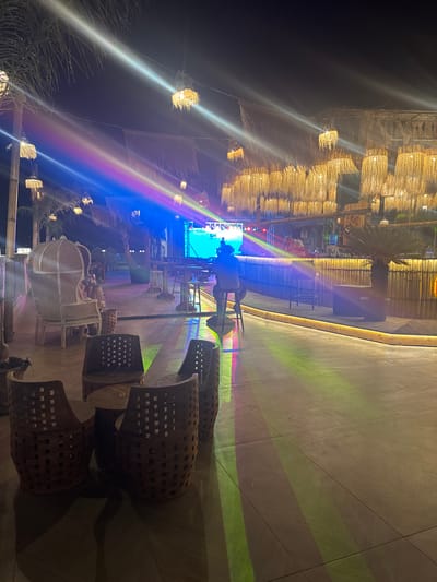 The image depicts a nighttime scene at a beachfront establishment in Golem, Albania. The establishment features a bar area with a large screen displaying vibrant colors, likely a visualizer or a performance. The bar itself is constructed from bamboo and is illuminated by lights. Several patrons are present; one person is seated at the bar, and another appears to be working behind it. Overhead, numerous lamps made of woven natural materials hang, casting a warm glow. The area is furnished with seating, including a distinctive armchair and woven chairs around small tables. The ground is paved with large tiles, and colorful light projections from the bar area create streaks of light across the surface. Palm trees and other tropical vegetation are visible, contributing to the ambiance. The overall impression is that of a lively, open-air venue with a tropical theme.