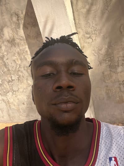 An adult male is depicted in a close-up, head-and-shoulders portrait, positioned against a textured wall in Birnin Kebbi, Nigeria. The subject has dark skin, short dark hair styled in braids or dreadlocks with faded sides, and a small goatee. He wears a basketball jersey with a split design; the left side (viewer's left) is predominantly dark brown with red and gold stripes, while the right side is white mesh featuring a red and blue NBA logo. His eyes are dark and appear slightly narrowed as he looks directly into the camera. The background consists of a light-colored, weathered concrete or plaster wall exhibiting discoloration and rough surface texture. A distinct shadow line from a vertical structural element (pillar or corner) is visible on the wall behind the subject's head. The scene is illuminated by bright natural light, casting clear shadows.