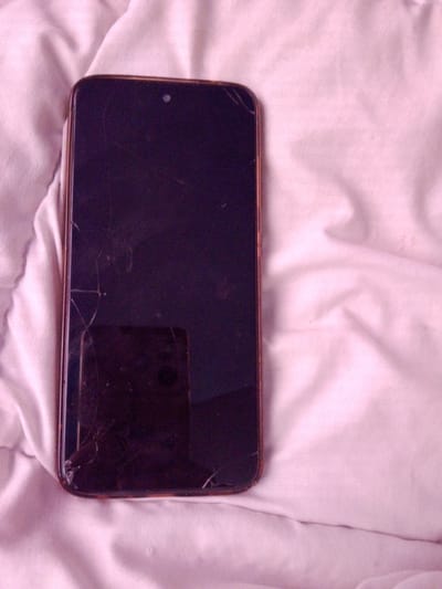 A dark-colored smartphone with a cracked screen lies on a rumpled pink fabric surface, possibly a blanket or bedding. The phone's screen is dark, and the casing appears to be a metallic brown. Visible cracks spiderweb across the glass. A reflection of the phone and what appears to be a ceiling light fixture is visible on the screen. The fabric surface is textured with folds and creases, indicating it is not flat. The image is taken from a slightly elevated angle, looking down at the phone. The location in the city of Inhulo-Kamianka, Ukraine is not discernible from the image itself.