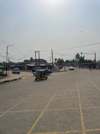 This image depicts a bustling intersection in Mosafejaw, Nigeria, under a bright, hazy sky. The foreground shows a grey interlocking brick road surface with prominent yellow cross-hatching, indicating a no-stopping zone.
Traffic is present, featuring a yellow motorized tricycle (keke napep) in the mid-ground, heading right, with at least two occupants. Behind it, a dark sedan is parked or stopped with its driver's side door open, adjacent to a white van also with an open door. Further left, a white pickup truck is partially visible, moving away from the viewer. In the distance, a white sedan approaches the intersection from the right. A large white truck is partially visible behind utility poles in the mid-ground.
Several pedestrians are visible; one person in a white top walks near the approaching white sedan, and two others are on the far right side of the road.
The intersection is lined with single-story buildings, many featuring corrugated metal roofs, indicative of commercial or residential structures. Dense utility poles with numerous overhead power lines crisscross the scene. A red traffic light is visible on the far right, and a road sign structure stands in the mid-ground. Palm trees and other tropical foliage are evident in the background, particularly on the right. A faint, irregular green line or streamer is visible in the upper left sky.