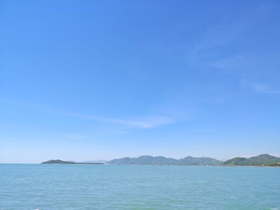 This image depicts a tranquil seascape with a clear blue sky and calm turquoise water. The horizon is lined with lush green mountains and a distant island, suggesting a coastal location. The water is gently rippling, reflecting the light of the sun. The sky is mostly clear, with a few wispy clouds scattered across it. The scene evokes a sense of peace and natural beauty. The image captures the coastal landscape of Ninh Ích, Vietnam, showcasing its picturesque coastline and mountainous backdrop.