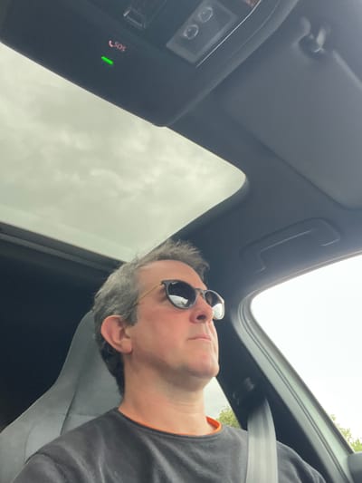 A middle-aged man with graying hair is visible in a car. He is wearing sunglasses and a seatbelt. The car has a sunroof that is open to a cloudy sky. A console with an "SOS" button and a green indicator light is visible. There is no verifiable evidence to indicate a location in the city of Bergheim, Germany.