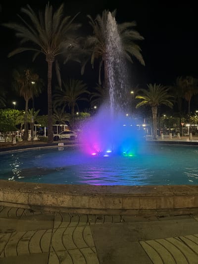 This media file depicts a nighttime scene at a public fountain in Sant Antoni de Portmany, Spain. The fountain features illuminated water jets, creating a vibrant display of blue and purple light. The water is illuminated from within, giving it an ethereal glow. Surrounding the fountain are several palm trees, their fronds softly lit by the ambient light. In the background, buildings with lights on are visible, suggesting an urban or resort setting. There are also some streetlights and outdoor seating areas with tables and chairs, indicating a public space that is open for leisure. The overall atmosphere is tranquil and visually appealing, suggesting a pleasant evening environment. The time of day is clearly night due to the dark sky and artificial lighting. There are no people clearly visible in the frame, but the presence of seating suggests a place where people might gather. The scene is likely outdoors, given the palm trees and the open-air fountain.