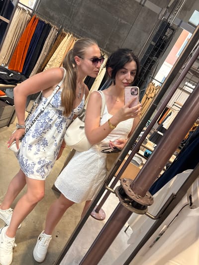 Two young women are taking a mirror selfie in what appears to be a clothing store. The woman on the left is wearing sunglasses, a white dress with a blue floral pattern, and white sneakers. She has her hair in a ponytail and is wearing a silver necklace and a watch. She is holding a white purse with a chain strap. The woman on the right is wearing a white dress with eyelet detailing and white sneakers. She has dark hair and is holding a pink phone in her hand, taking the picture. She is also wearing a gold watch and a delicate necklace. The background shows racks of clothing, some of which are colorful, and industrial-looking shelving and fixtures. The overall mood is casual and happy. The lighting suggests it is daytime.