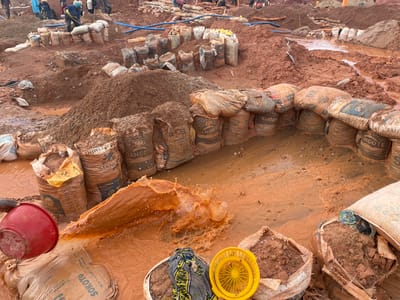 This media file captures a scene of what appears to be a mining or construction site in Rayfield, Nigeria, characterized by its muddy and wet terrain. The primary subjects are numerous sacks, many of which are filled with a brown, earthy substance and some appear to contain cement, indicated by visible text such as "DANGOTE," "SOKOTO," and "CEMENT." These sacks are arranged in piles and form a semi-circular barrier in the foreground, with some partially submerged in muddy water.

In the background, a group of people, dressed in casual clothing, are actively engaged in work. One individual is using a shovel, suggesting a process of excavation or material handling. Blue pipes are visible, possibly for water management or supply. The ground is a mixture of reddish-brown soil and mud, with puddles of water scattered throughout.

A red bucket is prominently featured in the foreground, from which a cascade of muddy water is being poured, creating a splash. Beside it, a yellow plastic basket and some fabric are placed on another sack. The overall impression is one of a busy, labor-intensive operation taking place in potentially challenging, wet conditions. The lighting suggests it could be daytime, though specific time or weather details are not discernible. The visible text on the sacks provides a strong clue to the location's context, linking it to cement production and distribution in Nigeria.