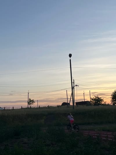 The image depicts a rural or suburban outdoor scene at dusk. A young person, appearing to be a child, is riding a bicycle on a reddish track in the foreground. The child is wearing a light-colored shirt and pink shorts. The surrounding environment is characterized by tall, dark green grass and vegetation. In the middle ground, there is a chain-link fence and sparse trees. In the background, there are power lines stretching across the frame, supported by utility poles. One prominent utility pole in the center of the image has a large, dark object attached to its top, possibly a siren or speaker. Beyond the power lines, the sky displays a gradient of colors, transitioning from a pale blue at the top to soft oranges and pinks near the horizon, indicative of sunset. Silhouetted structures, possibly rural buildings, are visible against the colorful sky. The overall impression is a quiet, possibly end-of-day scene in a somewhat undeveloped area. There is no specific information in the image to suggest the location is in the city of Koroviia, Ukraine.