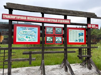 The image shows an outdoor informational display in Ust'-Pyzas, Russia. The display is mounted on a rustic wooden structure. The top sign, in red with white lettering, reads "ТРАНСПОРТИРОВКА ЛЕСОЗАГОТОВОК В ЛАГЕРНЫХ ОТДЕЛЕНИЯХ" which translates to "TRANSPORTATION OF LOGGING OPERATIONS IN CAMP SECTIONS". To the right of this sign is a smaller red sign with white numbers and text that reads "40-60 ГОДЫ", meaning "40s-60s YEARS".
Below the main sign, there are several framed exhibits. The leftmost exhibit is a map with the title "Схема УЖД лесоотделения п. Кантус (ВД 30/4)" which translates to "Map of the Narrow-Gauge Railway of the logging section of Kantus village (WD 30/4)". This map shows rivers, forests, settlements, and railway lines. To the right of the map are three framed photographs. The first photograph shows a snowy landscape with a river. The second photograph shows a narrow-gauge train locomotive. The third photograph shows people working in a snowy environment.
The display is set against a backdrop of green grass and trees, with a cloudy sky overhead. The wooden structure itself appears aged and weathered. Barbed wire is loosely wrapped around some of the wooden supports, adding to the rugged and possibly historical context of the display. The ground in front of the display is covered with gravel and some concrete. The scene suggests a historical or educational exhibit related to logging in the region.
