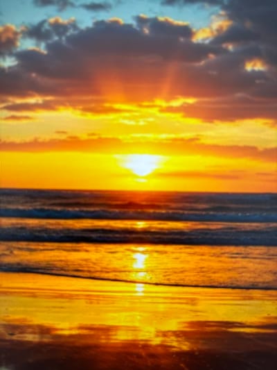 The image depicts a sunset over a body of water, likely the Ob River, given the context of Megion, Russia. The sky is a vibrant gradient of orange, yellow, and hints of blue, with scattered clouds tinged with the warm hues of the setting sun. Rays of light pierce through the clouds, casting a golden glow. The water reflects this fiery sky, creating a shimmering surface with distinct bands of light and shadow. The foreground shows a wet sandy or muddy shore, also reflecting the colors of the sky. There are no people or man-made objects visible in the scene.