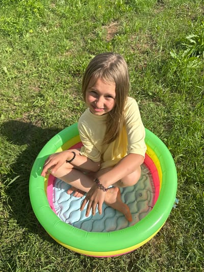 A young girl with blonde hair is sitting in a small, colorful inflatable pool in a grassy area. She is wearing a yellow t-shirt. The pool has green, yellow, and pink sections around its rim, and the interior is light blue with a quilted pattern. Her legs are bent, and her feet are visible inside the pool. She is looking towards the camera with a smile. Two dark bracelets are visible on her wrists. The scene appears to be outdoors on a sunny day, with green grass surrounding the pool. The sunlight casts shadows on the grass, suggesting it is midday. The presence of the inflatable pool and the sunny weather indicates a recreational activity, likely during warmer months. The location is identified as Mokino, Russia. There is no visible text in the image.
