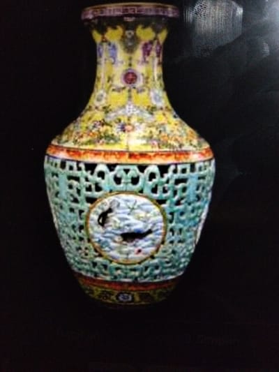 The image shows a close-up, low-resolution photograph of an ornate porcelain vase against a dark, possibly black, background. The vase is decorated with intricate patterns and imagery. The upper portion of the vase is predominantly yellow with multi-colored floral and figural designs. A red circular motif is visible on the neck of the vase. The middle section of the vase features a celadon-green, openwork lattice design. Within the lattice, a circular panel displays a scene with water and a dark bird-like figure. The base of the vase has a band of red and yellow decoration. The lighting appears to be from above and slightly to the right, casting some subtle shadows. The image quality is poor, with visible noise and blur, particularly around the edges and in darker areas. There is no discernible environmental context suggesting a location in Kualaraja, Indonesia, or any other specific urban setting. The image solely focuses on the object itself.