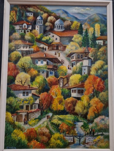 An oil painting depicts a stylized, elevated view of the city of Elena, Bulgaria. The artwork, framed in white, showcases the town nestled into a hillside with a mountain range in the distance. Buildings with white walls and red-tiled roofs are surrounded by dense, colorful trees exhibiting autumnal hues of yellow, orange, and green. Several figures are visible walking along pathways, and a small bridge spans a narrow body of water. A church with a white dome and a small number of windows is situated on the higher part of the city. In the lower right corner of the painting, there is a signature that reads "C. CABOB."