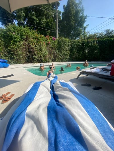 A sunny outdoor scene features a pool with several people swimming and socializing. The pool is bordered by lush greenery and a stone patio area. A large blue and white striped towel dominates the foreground, suggesting a relaxed poolside setting. A pair of sandals sits nearby. In the distance, power lines crisscross the sky.