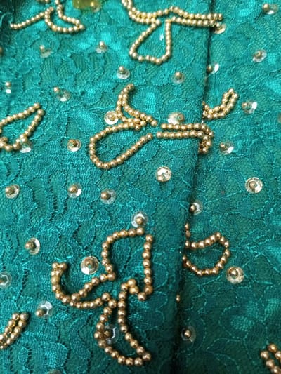 A textured turquoise fabric, possibly lace, displays embellishments. Shiny, gold-colored beads trace intricate floral or abstract patterns. Small, clear, flat sequins or beads are scattered sparsely across the background, enhancing the overall decorative effect. The texture of the fabric has noticeable variation.