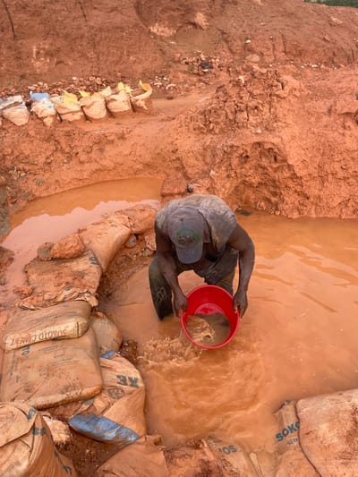 This overhead shot captures a person engaged in a manual labor activity in a muddy, outdoor setting. The individual, wearing a grey t-shirt, dark pants, and a blue baseball cap, is in a pool of muddy water, holding a bright red basin. They appear to be panning for materials within the basin, suggesting a process of sifting or washing.

The environment is characterized by red-brown earth and large, burlap sacks that are partially submerged in the water or stacked along the muddy banks. Some of these sacks are clearly labeled with "SOKOTO CEMENT" and "42.5N," indicating a potential connection to construction or materials processing. The water is a murky, reddish-brown color, suggesting that the soil is rich in iron oxides.

The scene appears to be a work site, possibly related to mining or resource extraction, given the presence of the water, mud, and sacks of cement. The lighting suggests it is daytime, and the activity implies arduous physical work. There are no explicit cues about the time of day or weather, beyond the muddy conditions which could be a result of recent rain or the ongoing activity. The overall impression is one of a rugged, practical operation.