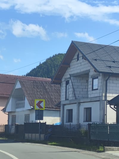 The image shows a street scene in Leordina, Romania, with several houses visible. The foreground features a paved road with white lane markings. Behind the road, there is a dark metal fence and grass. Several houses are depicted: one on the left with a pinkish facade and a brown tiled roof, a white house with a corrugated roof in the center, and a house under construction on the right, made of white cinder blocks with a dark tiled roof. A yellow and red chevron traffic sign indicating a curve to the left is mounted on a pole in front of the central white house. A person is seated on a bench behind the fence, facing away from the camera, wearing a plaid shirt and blue jeans. The background consists of a tree-covered hillside under a bright blue sky with scattered white clouds. Power lines are visible crisscrossing the sky.