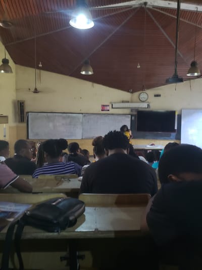 The image depicts an indoor scene, likely a classroom or lecture hall, with a group of people seated at desks, facing a presenter. The ceiling is constructed of wooden planks with exposed beams and features several industrial-style light fixtures. A large, dark screen is mounted on the wall behind the presenter. Two whiteboards are also visible on the wall. The overall atmosphere appears to be that of an educational setting. The image is likely taken in Iganmu, Nigeria, given the context of the prompt.