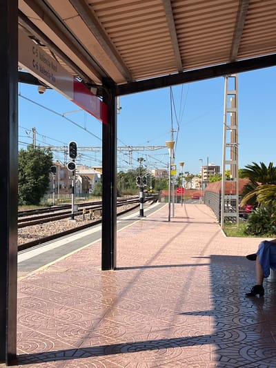 The image shows a train station platform in Puçol, Spain. A sign reads "C-5 Valencia-Nord, C-6 Valencia-Nord" indicating train lines. The platform surface has a patterned tile design. Rail tracks run alongside the platform. A person's legs and feet, wearing a blue dress and black shoes, are partially visible at the edge of the platform. Overhead power lines stretch above the tracks. Distant buildings and greenery are visible. A covered structure provides shelter on the platform. Signal lights are visible along the tracks.