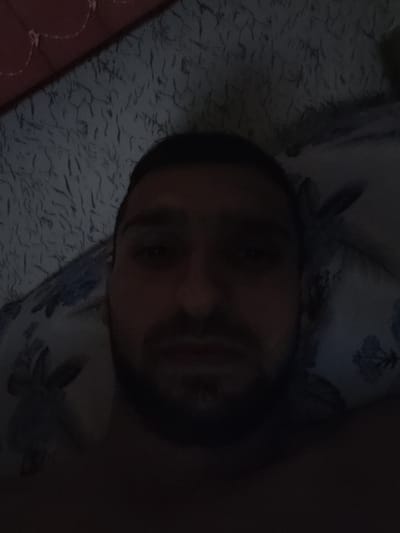 The image is a dark, low-resolution selfie taken from a close-up perspective. A man's face is partially visible, looking directly at the camera. His facial features are obscured by shadow, though it appears he has dark hair and a beard. He is lying down, and the background consists of a textured surface, likely a bed or pillow, with a patterned fabric visible to his right and a reddish, textured material to his left, possibly a blanket or headboard. The overall lighting is dim, suggesting the photo was taken indoors in low light conditions. No specific location in Pirdop, Bulgaria is discernible from the visual information.