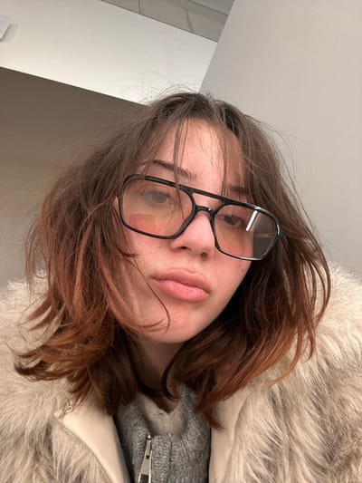A female individual is depicted in a close-up, chest-up portrait shot, reportedly captured in Reichstett, France. She presents with shoulder-length, disheveled brown hair displaying reddish highlights, particularly towards the ends. The subject wears large, black-framed eyeglasses with lightly tinted lenses. Her mouth is slightly open, forming a subtle pout. She is dressed in a light-colored, possibly off-white or cream, faux fur jacket featuring a distinct, smooth collar element. Underneath the jacket, a grey knitted garment with a visible zipper mechanism is present. The background is a plain, light-toned wall or ceiling surface, indicating an indoor environment. The individual's gaze is directed towards the camera.